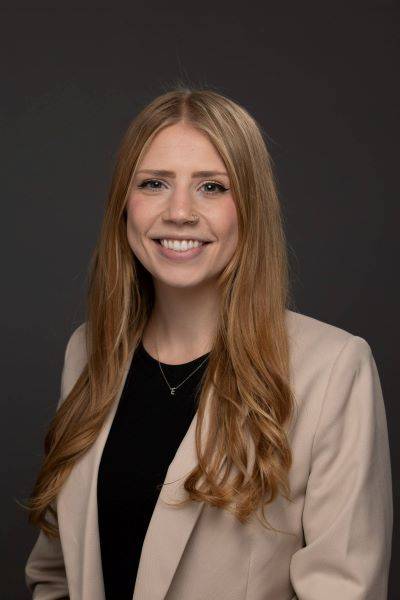 A formal headshot of Elizabeth Clark who has long blonde hair and is wearing a tan suit jacket and a black blouse